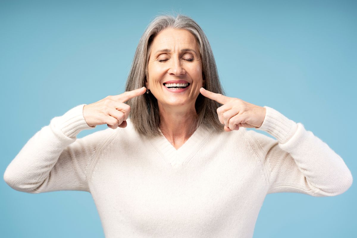 Older woman pointing to her teeth while smiling, representing dental care and tooth replacement.